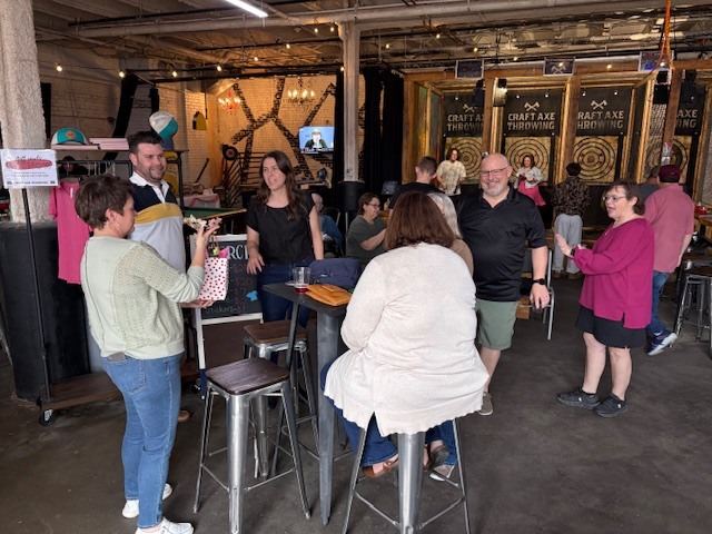 Guests gathered around high-top tables inside Craft Axe Throwing Spartanburg, with the axe-throwing lanes behind them