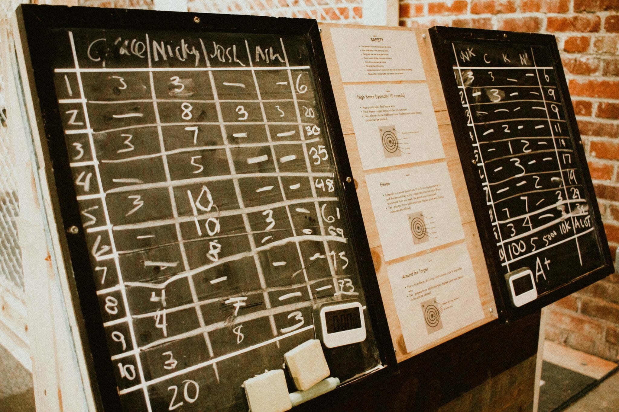 Two chalkboard scoreboards at Craft Axe Throwing showing ten rounds of scores for four players, beside printed cards explaining the safety rules and scoring games