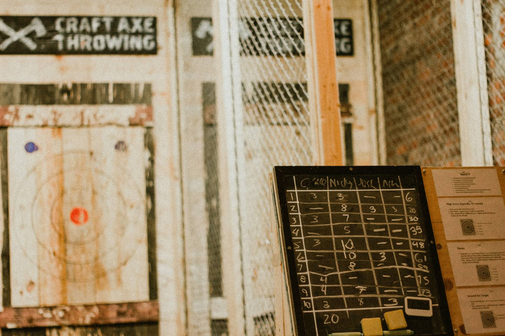 Enclosed axe throwing lanes at Craft Axe Throwing with mesh safety fencing, a wooden bullseye target, a chalk scoreboard, and posted safety rules