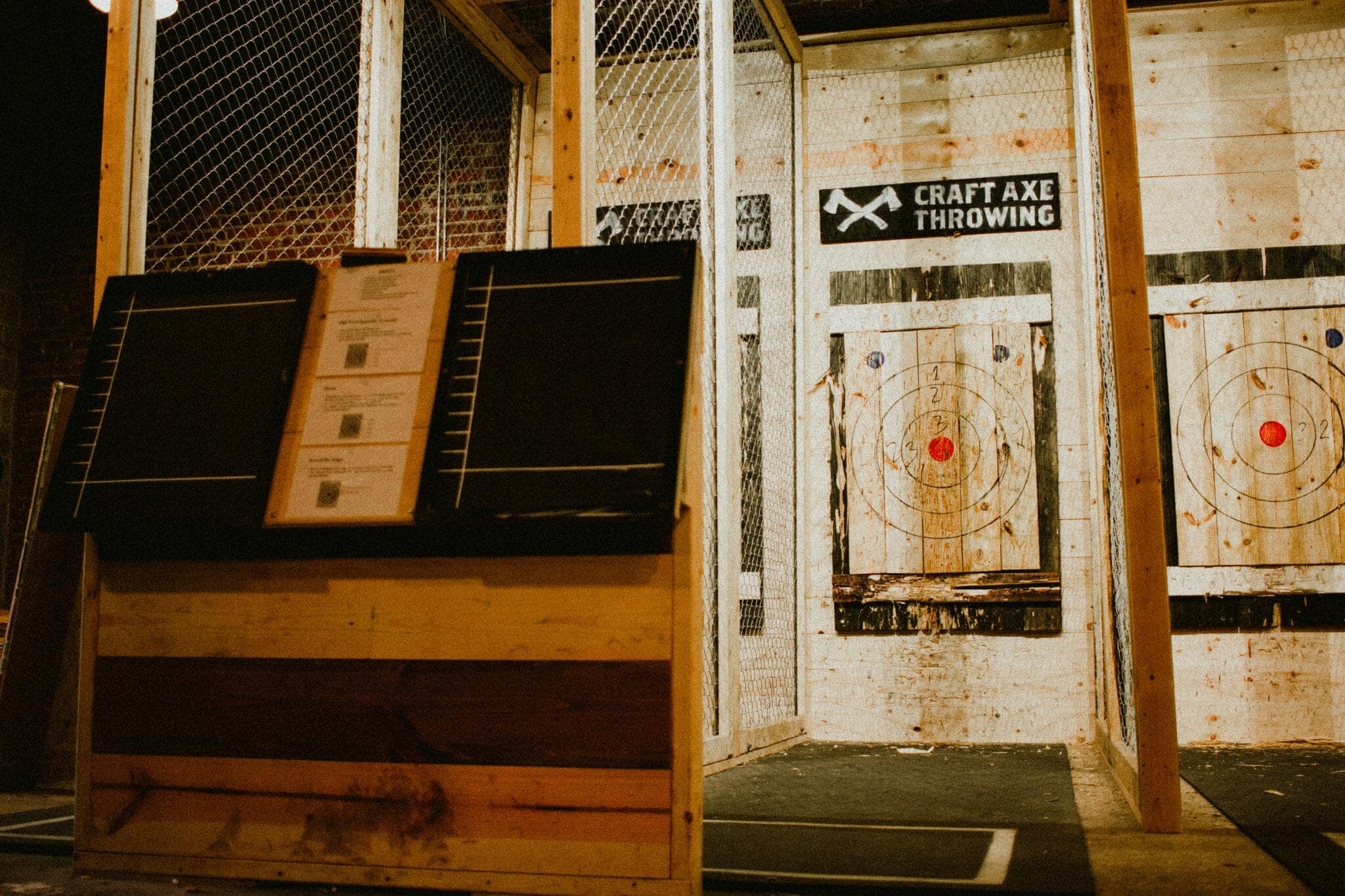 Enclosed axe throwing lanes at Craft Axe Throwing, each caged in wood and chain-link mesh with wooden bullseye targets and posted safety rules under the Craft Axe Throwing logo