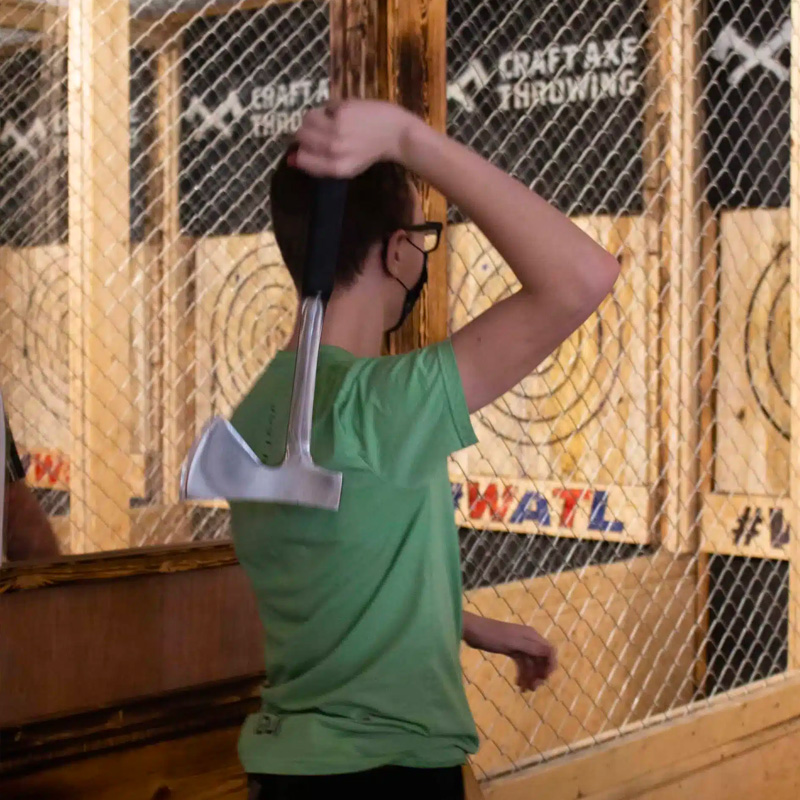 A thrower with an axe raised overhead mid-windup, lining up a throw at a wooden bullseye target in an enclosed axe throwing lane