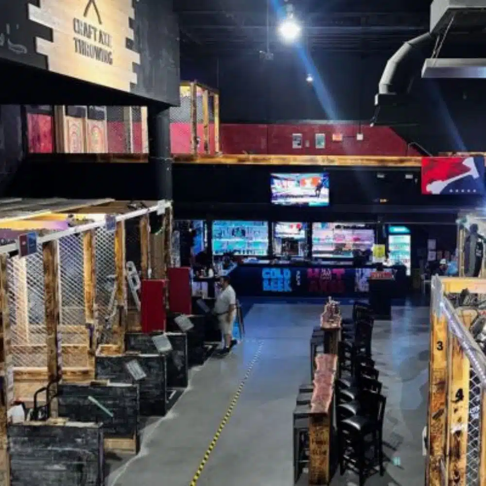 Row of caged axe throwing lanes leading toward a bar with TVs and cold-beer-hot-axes signage inside a Craft Axe Throwing venue